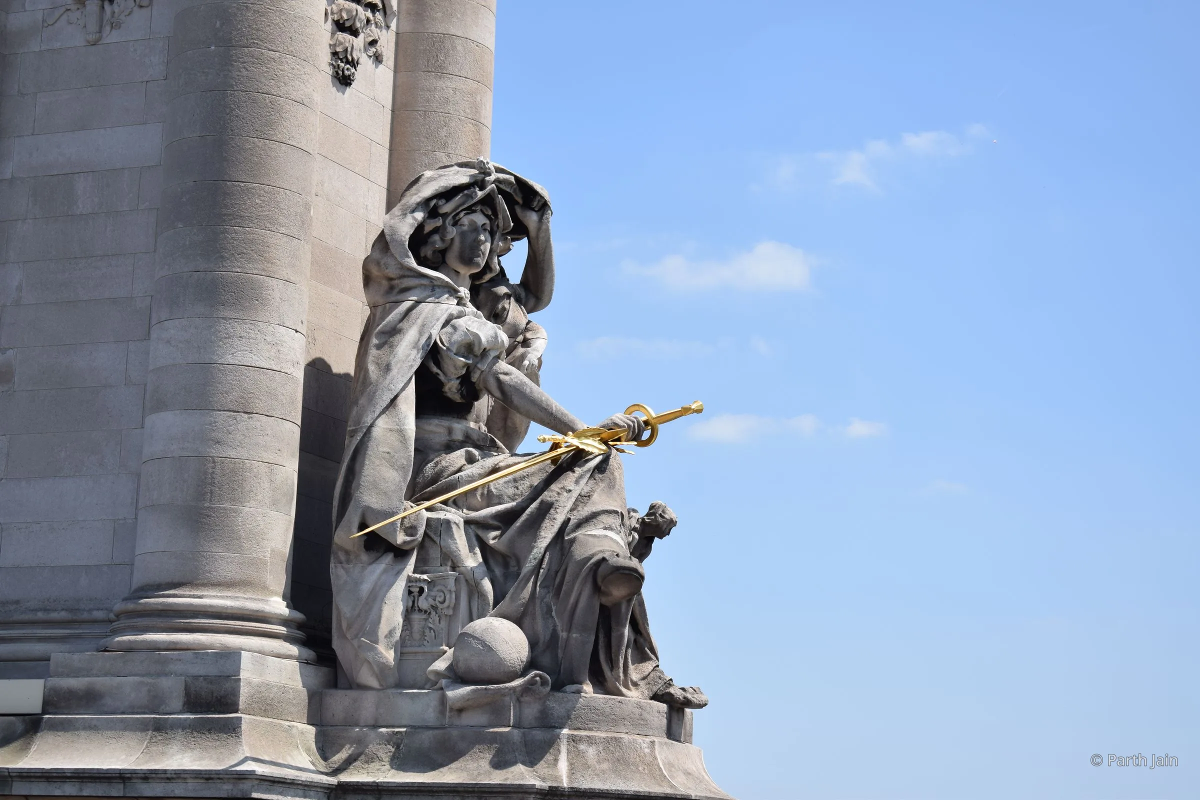 A stone allegorical figure on the Pont Alexandre III pylon, holding a gilded sword and shield against a blue sky.