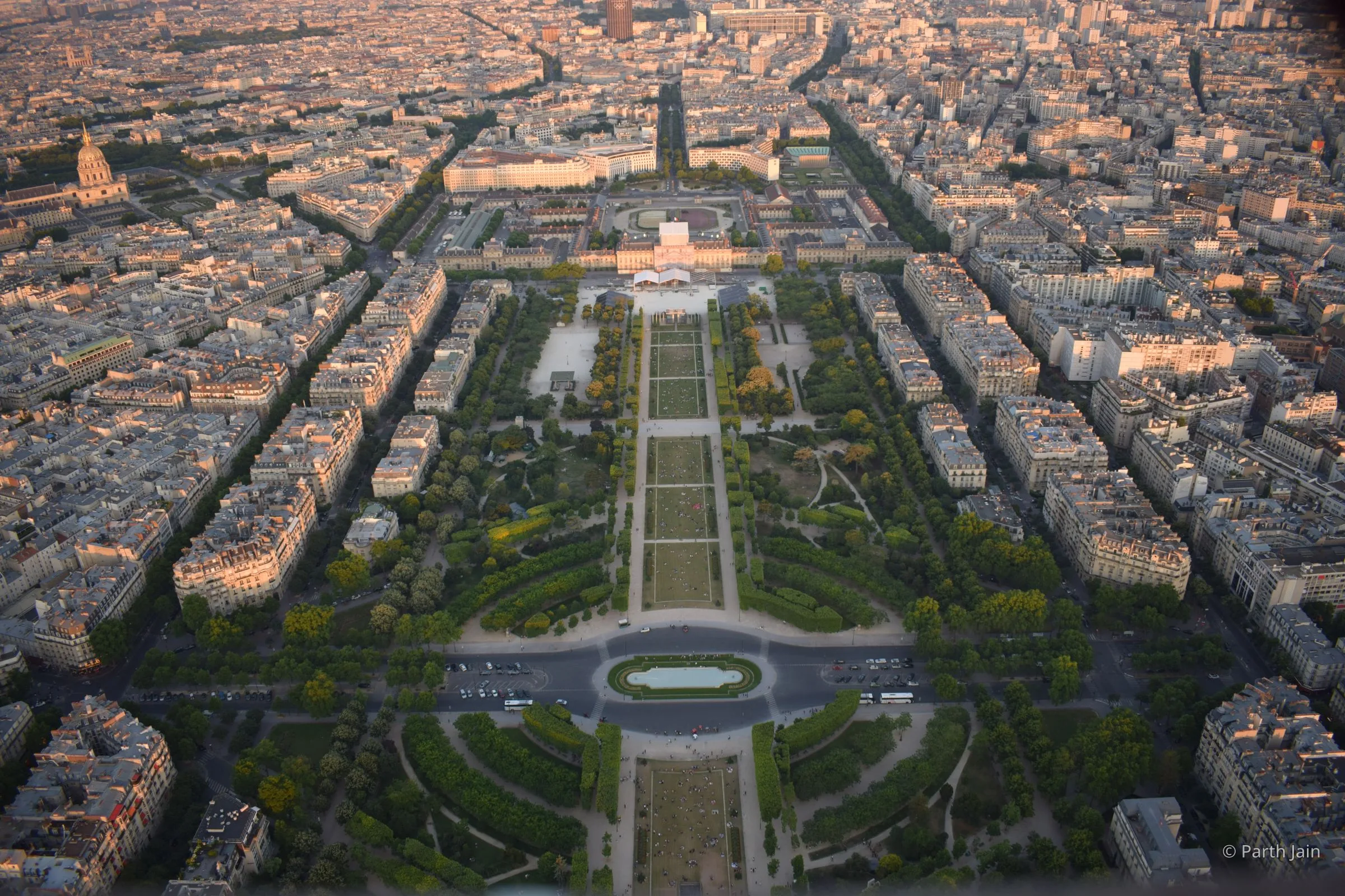 Aerial view of the Champ de Mars from the top of the Eiffel Tower at golden hour, École Militaire centred at the far end and Les Invalides golden dome on the upper-left horizon.